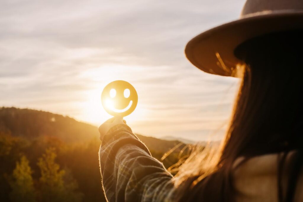 person standing in rugged outdoor setting at sunrise holding up a cut-out of a smiley face so that the light shines through and they experience the 5 benefits of wilderness therapy