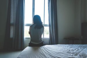 Young woman seated on her bed alone in her room staring out her window and wondering what are trauma triggers.