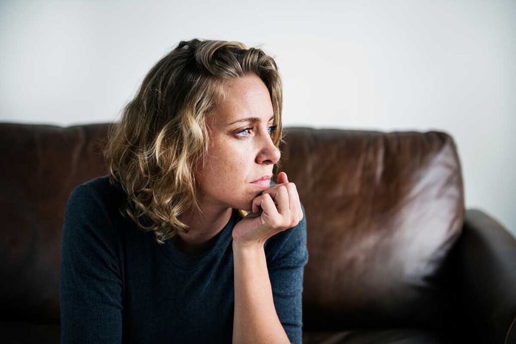 Woman seated on couch in an office setting and looking pensive as she ponders, "Can you be addicted to painkillers?"