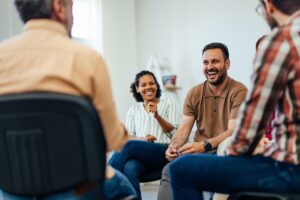 A group of adults sitting in a circle during a therapy session, smiling and engaging in conversation.