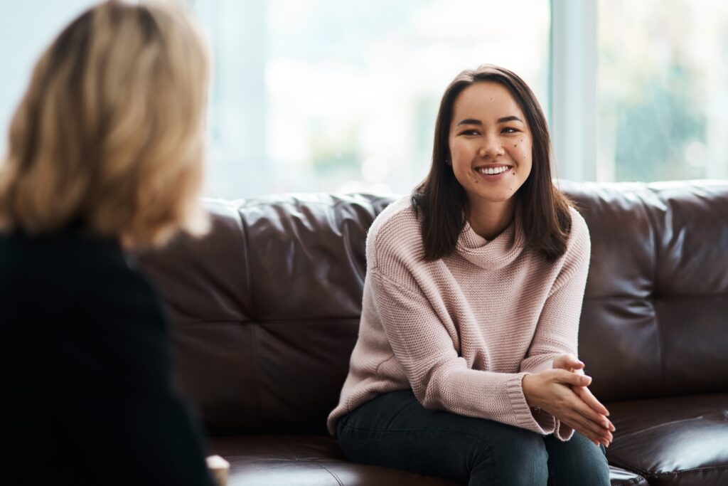 A smiling woman in a light pink sweater sits on a brown leather couch, leaning slightly forward while talking with another person whose back is facing the camera.